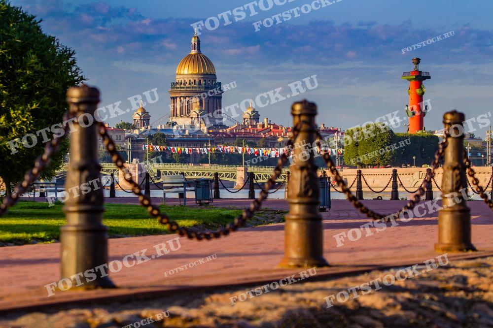 На фотографии представлен вид на Исаакиевский собор и Ростральные колонны в Санкт-Петербурге. Снимок выполнен с низкой точки съёмки через цепное ограждение, которое создаёт красивую перспективу и направляет взгляд на архитектурные символы города. Время суток — вечер или утро, что видно по мягкому и рассеянному свету, придающему сцене умиротворённый вид. Это изображение подходит для иллюстрирования путеводителей, исторических книг, культурных статей, а также может использоваться для украшения интерьеров и в качестве фотообоев.